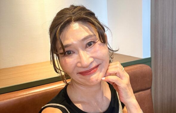 黒いトップスとシルバーのキャミソールを重ね、笑顔で椅子に座る女性