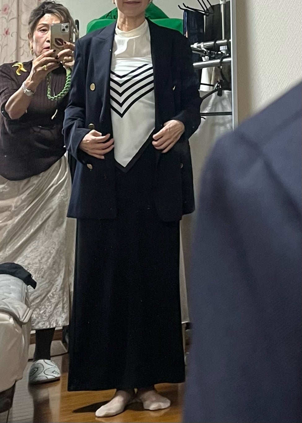 ジャケットと白いトップス、黒いラインのスカーフを合わせ、鏡で装いを確認する様子